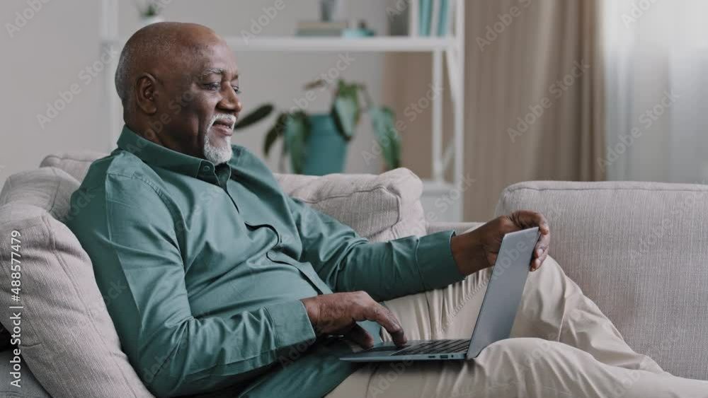 Happy mature pensioner sitting on sofa making internet order using modern laptop african american shopper browsing website smiling carefree elderly grandpa pays bank bills remote communication concept Happy mature pensioner sitting on sofa making internet order using modern laptop african american shopper browsing website smiling carefree elderly grandpa pays bank bills remote communication concept