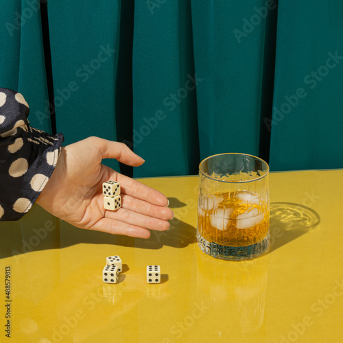 Creative arrangement made of playing dices, a glass and a woman's hand on a yellow table and green curtains. Minimal drinking concept. Party, gambling and elegance inspiration.