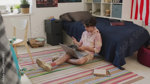 Long shot of curly-haired Caucasian male college student wearing hoodie, sitting on floor in dorm room at daytime, using portable computer and writing in notebook photo