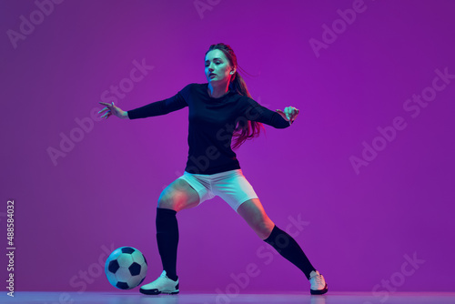 Professional female soccer player dribbling football ball isolated on purple studio background in neon light. Sport, action, motion, fitness