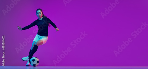 Professional female soccer player dribbling football ball isolated on purple studio background in neon light. Sport, action, motion, fitness