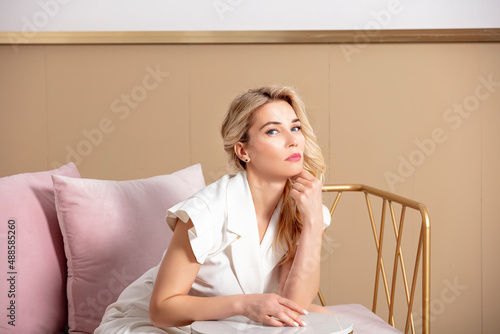 Portrait of a beautiful blonde woman in a white jacket dress sitting on a sofa in a stylish interior