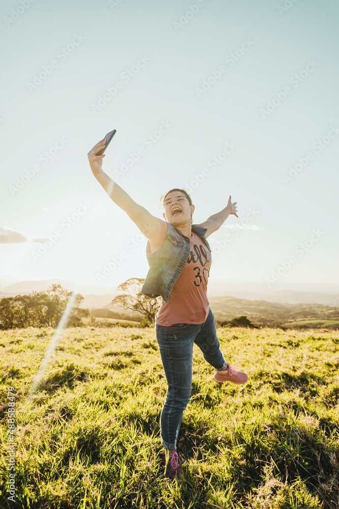 Vertical of a beautiful Caucasian woman outdoors taking a picture with her cell phone jumping very happy in a sunset. Vertical of a beautiful Caucasian woman outdoors taking a picture with her cell phone jumping very happy in a sunset.