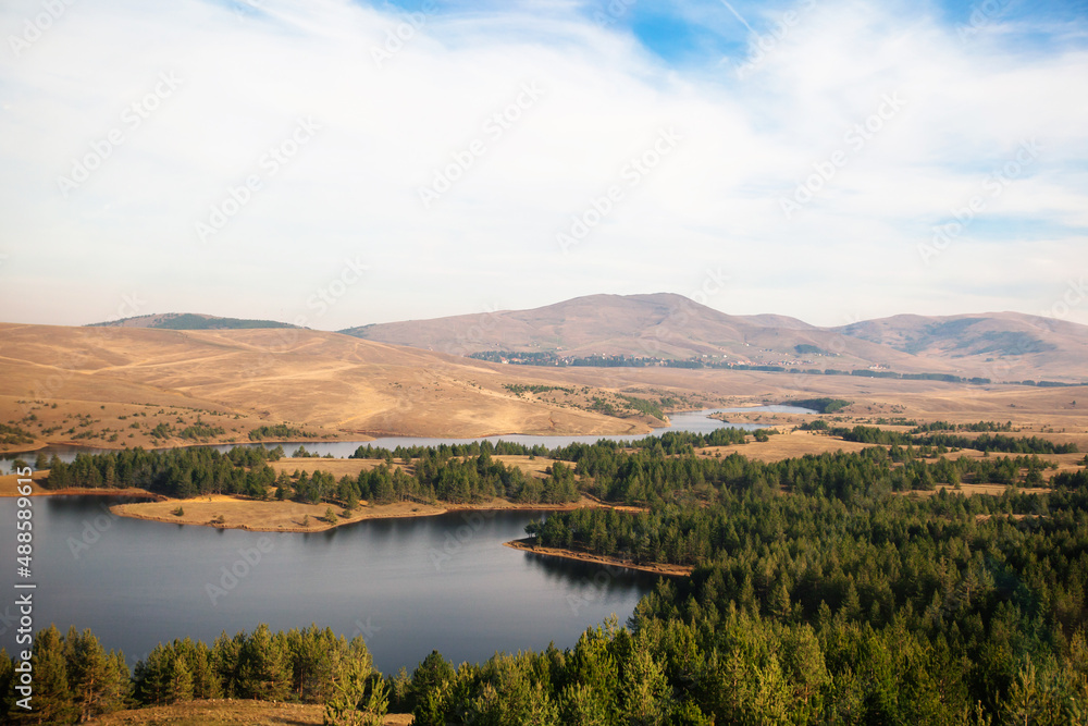 Autumn nature lake landscape view, hills and fields,   beautiful sky in background. Pine trees at lakes shore.