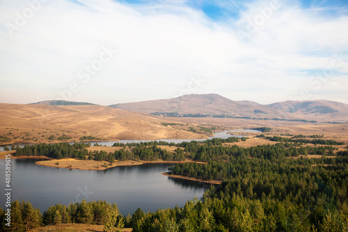 Autumn nature lake landscape view, hills and fields, beautiful sky in background. Pine trees at lakes shore.