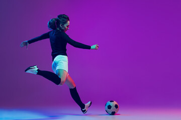 Dynamic portrait of female soccer player practicing with football ball isolated on purple studio background in neon light. Sport, action, motion, fitness