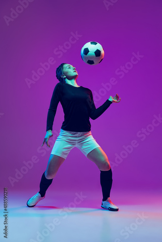 Emotive girl, female soccer, football player in motion, action isolated on purple studio background in neon light. Sport, active, healthy lifestyle