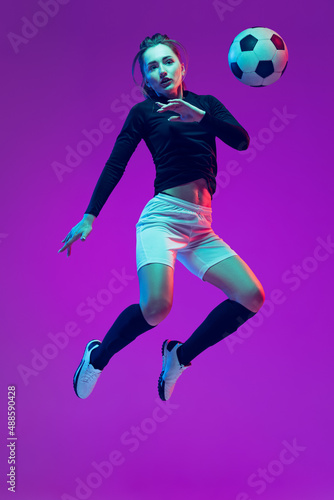 In action. One sportive girl, female soccer player training with football ball isolated on purple studio background in neon light. Sport, action, motion, fitness