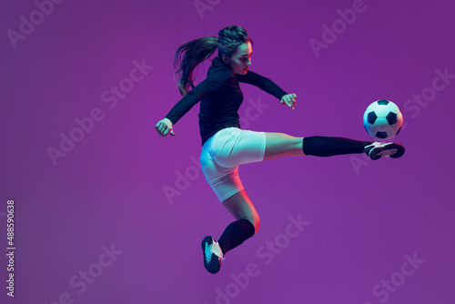 In action. One sportive girl, female soccer player training with football ball isolated on purple studio background in neon light. Sport, action, motion, fitness