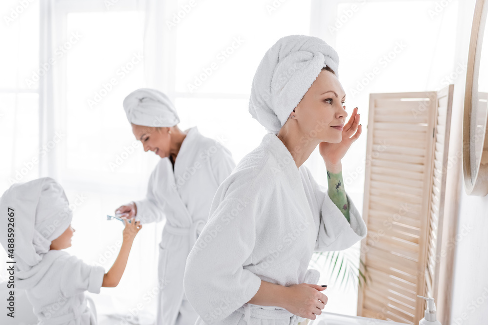 Lesbian woman in bathrobe applying cream near blurred girlfriend and adopted daughter at home