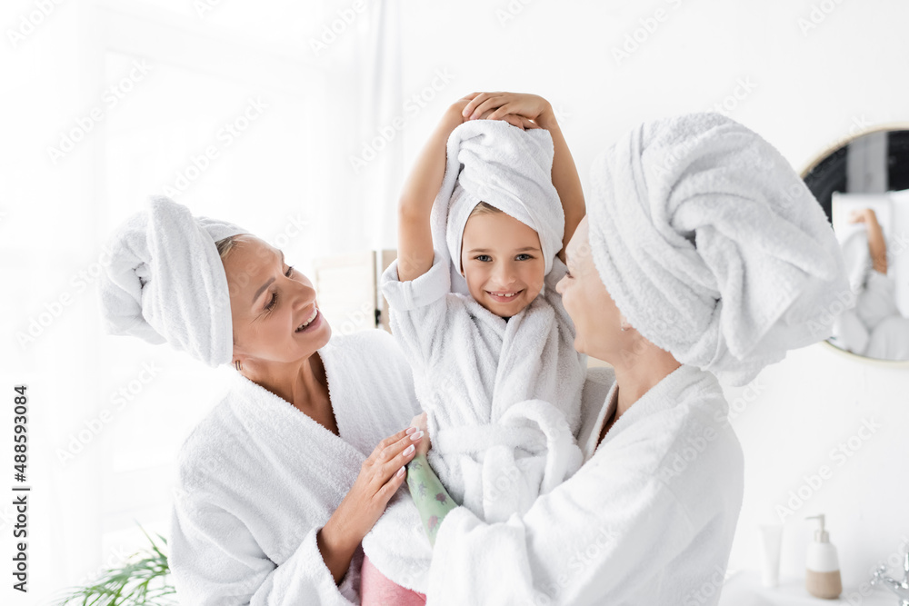 Cheerful lesbian women in bathrobes holding child in towel in bathroom Cheerful lesbian women in bathrobes holding child in towel in bathroom