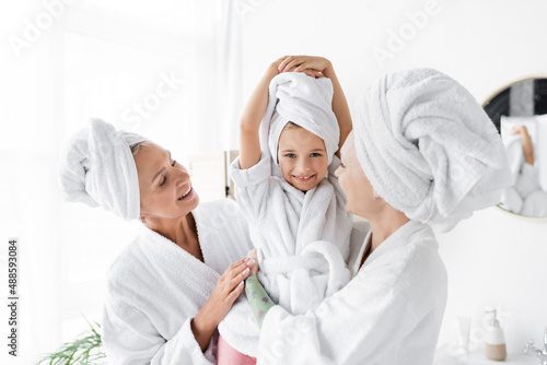 Cheerful lesbian women in bathrobes holding child in towel in bathroom