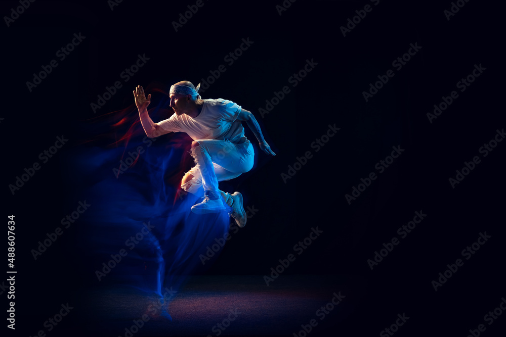 Fototapeta premium Jumping. Stylish man in sports white outfit dancing hip-hop, breakdance isolated on dark background in mixed neon light. Youth culture, hip-hop, style and fashion, action.