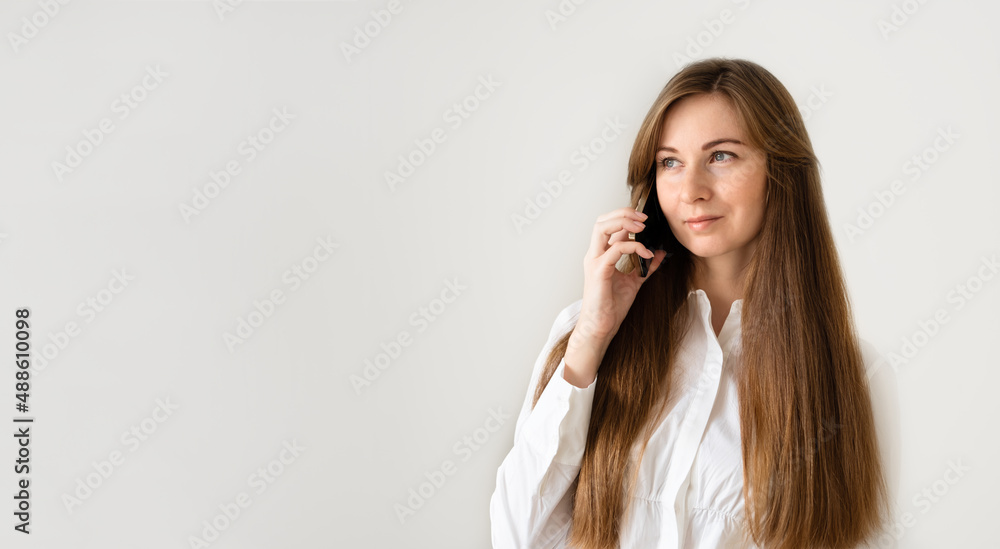 Caucasian woman with long brown hair smiling, speaking by smartphone,having call on phone on white,grey background at home or office.Banner,copy space Caucasian woman with long brown hair smiling, speaking by smartphone,having call on phone on white,grey background at home or office.Banner,copy space