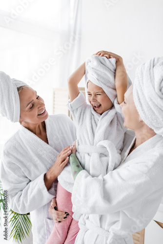 Happy lesbian women in bathrobes holding positive adopted daughter in bathroom