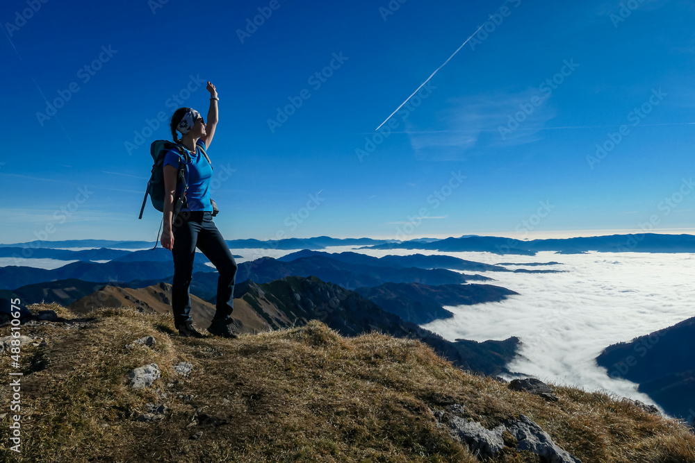Obraz premium Woman with backpack hiking and admiring a scenic view from mount Eisenerzer Reichenstein in Styria, Austria, Europe. Ennstal valley is covered in clouds. Hiking trail, Wanderlust. Freedom concept