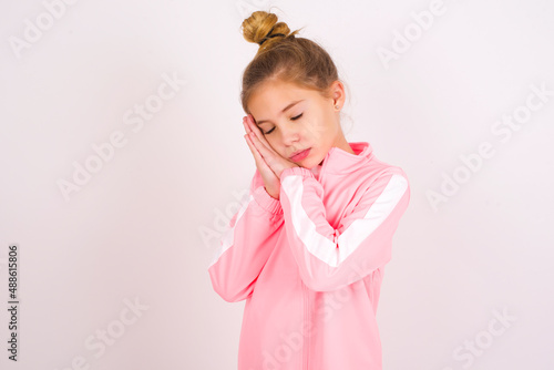 caucasian little kid girl with bun hairstyle wearing pink tracksuit over white background sleeping tired dreaming and posing with hands together while smiling with closed eyes.