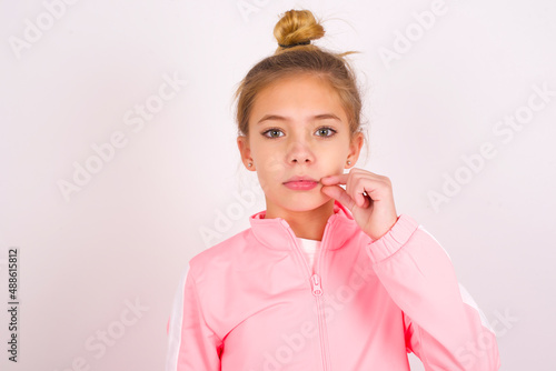 caucasian little kid girl with bun hairstyle wearing pink tracksuit over white background mouth and lips shut as zip with fingers. Secret and silent, taboo talking.