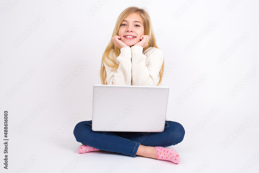 Dreamy caucasian teen girl sitting with laptop in lotus position on white background keeps hands pressed together under chin, looks with happy expression, has toothy smile. Dreamy caucasian teen girl sitting with laptop in lotus position on white background keeps hands pressed together under chin, looks with happy expression, has toothy smile.