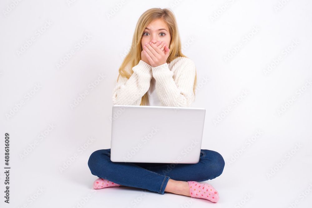 Upset caucasian teen girl sitting with laptop in lotus position on white background , covering her mouth with both palms to prevent screaming sound, after seeing or hearing something bad. Upset caucasian teen girl sitting with laptop in lotus position on white background , covering her mouth with both palms to prevent screaming sound, after seeing or hearing something bad.