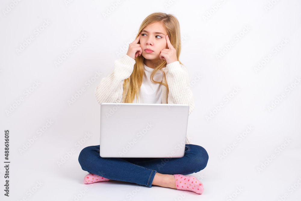 caucasian teen girl sitting with laptop in lotus position on white background with thoughtful expression, looks away, keeps hand near face, thinks about something pleasant. caucasian teen girl sitting with laptop in lotus position on white background with thoughtful expression, looks away, keeps hand near face, thinks about something pleasant.