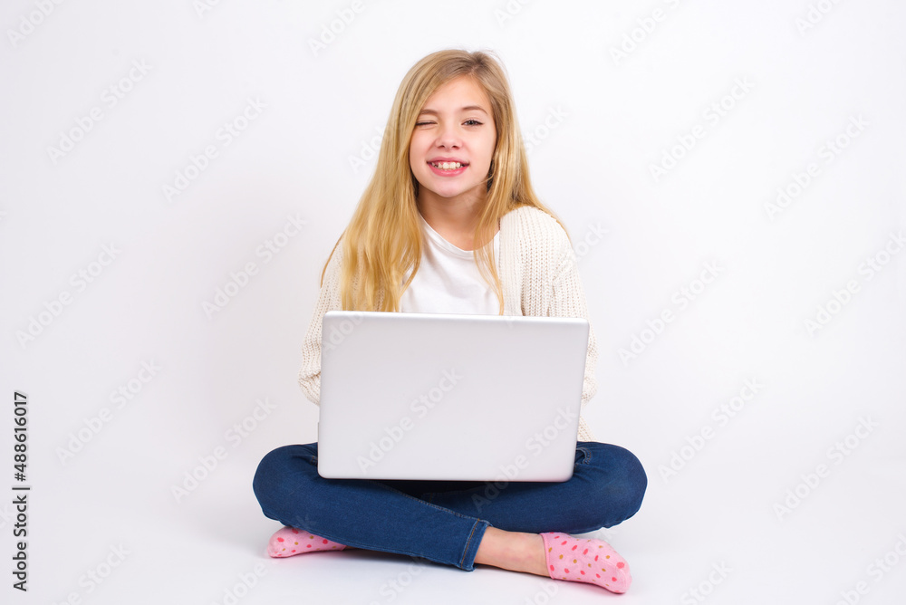 caucasian teen girl sitting with laptop in lotus position on white background blinking eyes with pleasure having happy expression. Facial expressions and people emotions concept. caucasian teen girl sitting with laptop in lotus position on white background blinking eyes with pleasure having happy expression. Facial expressions and people emotions concept.