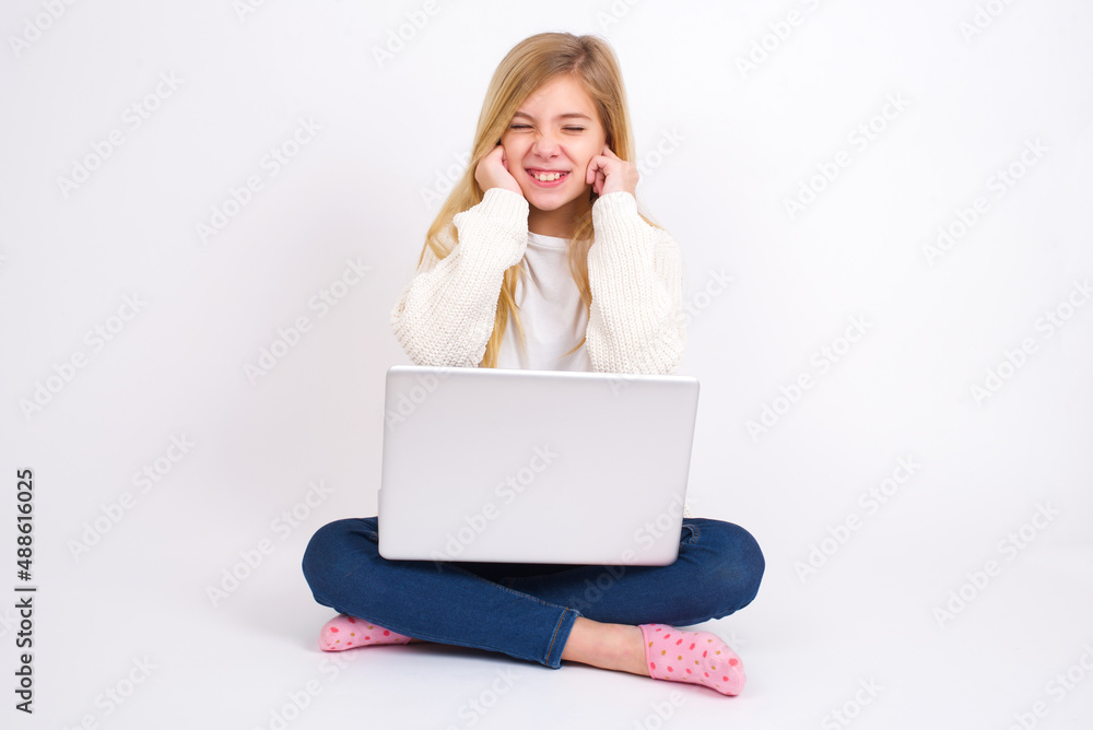 Stop making this annoying sound! Unhappy stressed out caucasian teen girl sitting with laptop in lotus position on white wall making worry face, plugging ears with fingers, irritated with loud noise. Stop making this annoying sound! Unhappy stressed out caucasian teen girl sitting with laptop in lotus position on white wall making worry face, plugging ears with fingers, irritated with loud noise.