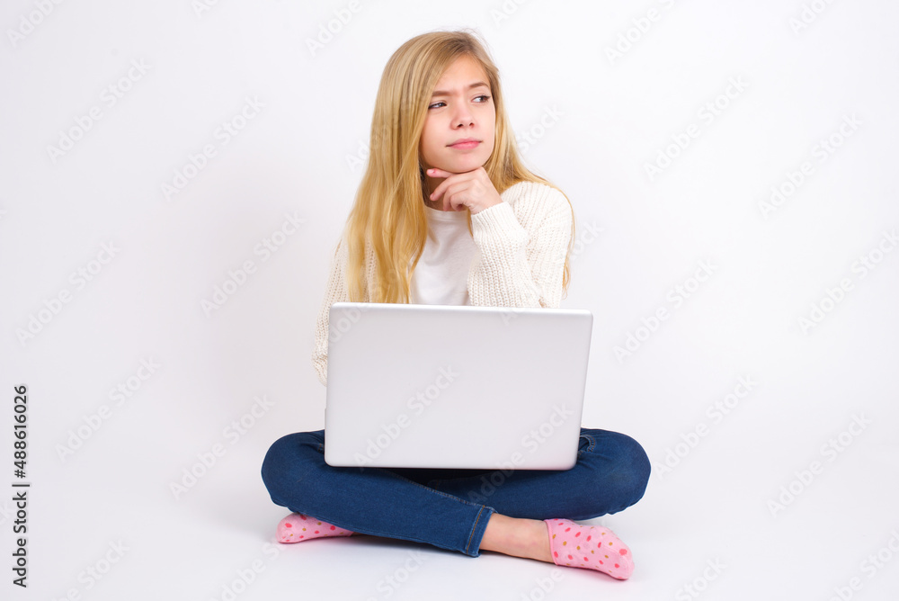 Dreamy caucasian teen girl sitting with laptop in lotus position on white background with pleasant expression, looks sideways, keeps hand under chin, thinks about something pleasant.