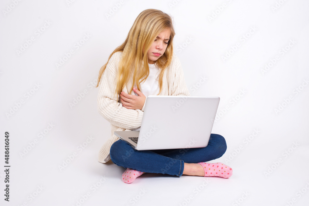 caucasian teen girl sitting with laptop in lotus position on white background suffering from strong stomachache. caucasian teen girl sitting with laptop in lotus position on white background suffering from strong stomachache.