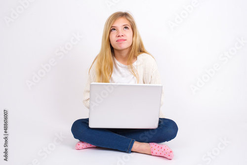 caucasian teen girl sitting with laptop in lotus position on white background has worried face looking up lips together, being upset thinking about something important, keeps hands down. © Jihan