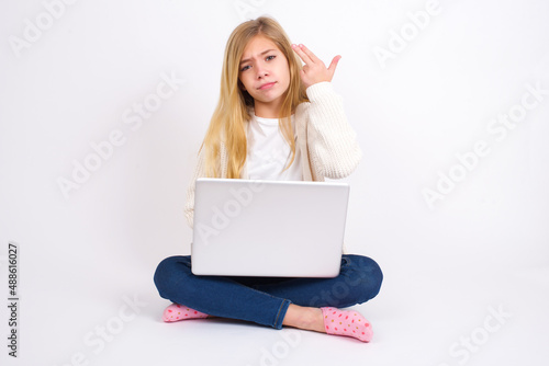 Unhappy caucasian teen girl sitting with laptop in lotus position on white background imitates gun shoot makes suicide gesture keeps two fingers on temples. © Jihan