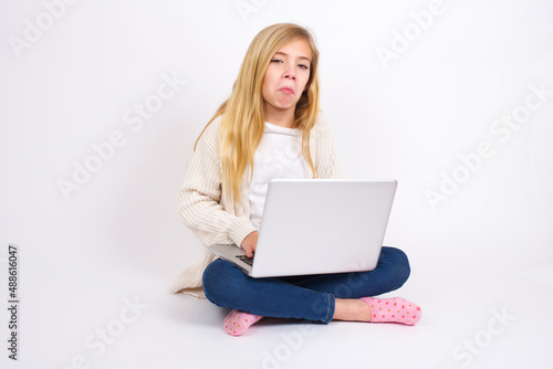 caucasian teen girl sitting with laptop in lotus position on white wall with snobbish expression curving lips and raising eyebrows, looking with doubtful and skeptical expression, suspect and doubt © Jihan
