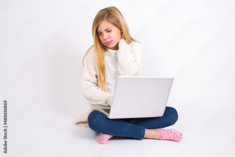 caucasian teen girl sitting with laptop in lotus position on white background suffering from back and neck ache injury, touching neck with hand, muscular pain. caucasian teen girl sitting with laptop in lotus position on white background suffering from back and neck ache injury, touching neck with hand, muscular pain.