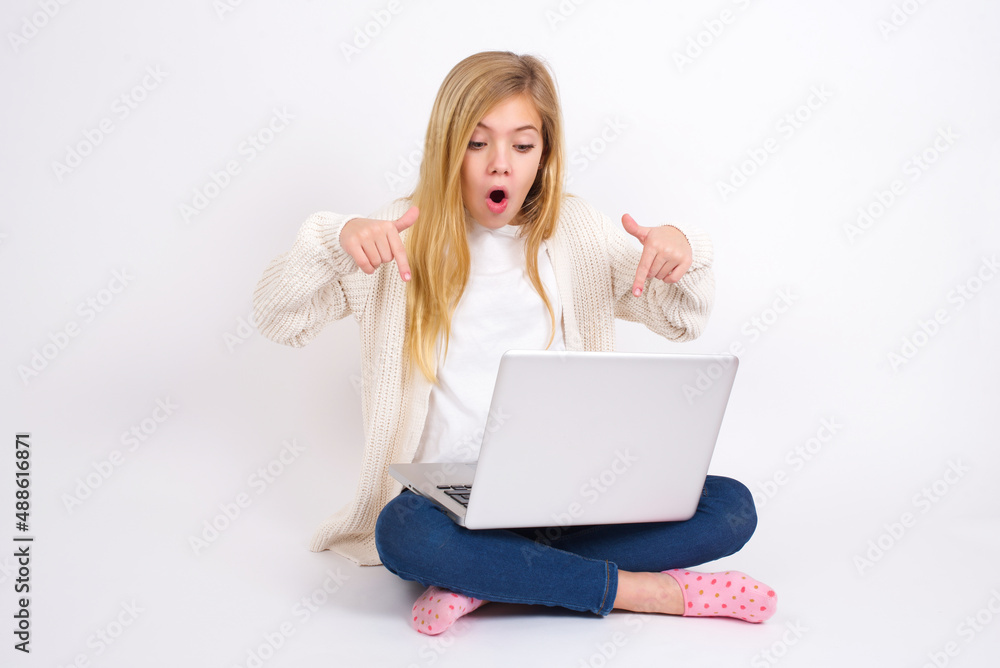 Naklejka premium Amazed caucasian teen girl sitting with laptop in lotus position on white background points down with fore fingers, opens mouth being shocked. Advertisement concept.