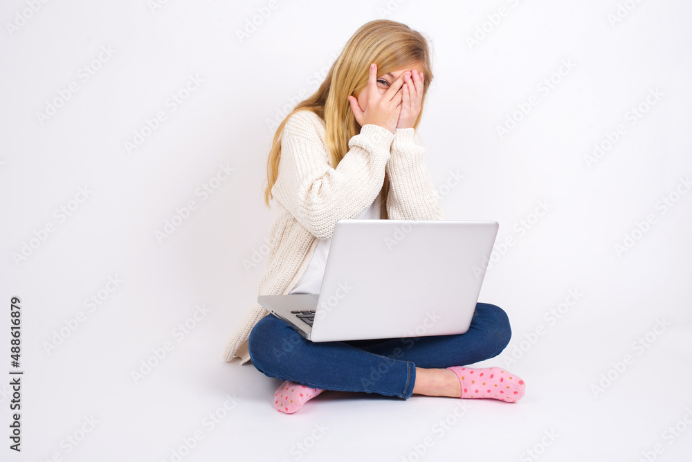 caucasian teen girl sitting with laptop in lotus position on white background covering face with hands and peering out with one eye between fingers. Scared from something or someone. caucasian teen girl sitting with laptop in lotus position on white background covering face with hands and peering out with one eye between fingers. Scared from something or someone.