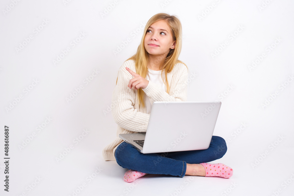 Portrait of caucasian teen girl sitting with laptop in lotus position on white background posing on camera with tricky look, presenting product with index finger. Advertisement concept. Portrait of caucasian teen girl sitting with laptop in lotus position on white background posing on camera with tricky look, presenting product with index finger. Advertisement concept.