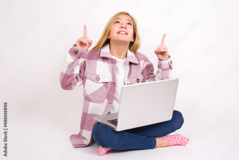 Successful friendly looking caucasian teen girl sitting with laptop in lotus position on white background exclaiming excitedly, pointing both index fingers up, indicating something. Successful friendly looking caucasian teen girl sitting with laptop in lotus position on white background exclaiming excitedly, pointing both index fingers up, indicating something.