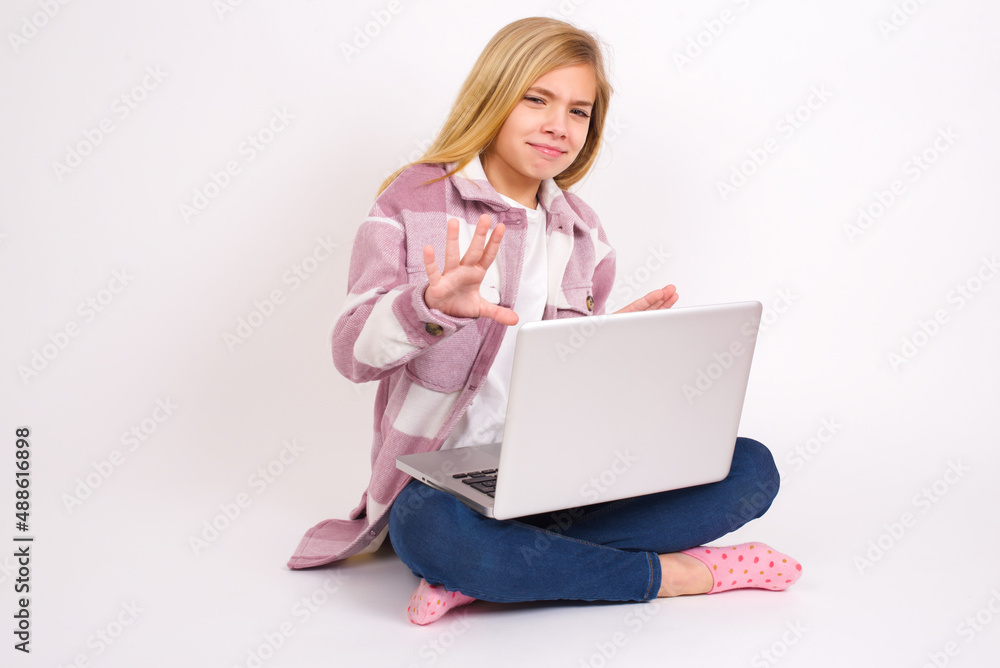 Afraid caucasian teen girl sitting with laptop in lotus position on white background , makes terrified expression and stop gesture with both hands saying: Stay there. Panic concept. Afraid caucasian teen girl sitting with laptop in lotus position on white background , makes terrified expression and stop gesture with both hands saying: Stay there. Panic concept.