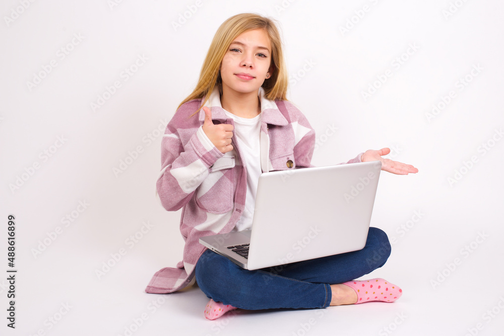 Happy cheerful caucasian teen girl sitting with laptop in lotus position on white background showing thumb up and pointing with the other hand. I recommend this. Happy cheerful caucasian teen girl sitting with laptop in lotus position on white background showing thumb up and pointing with the other hand. I recommend this.