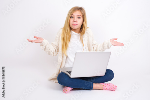 Puzzled and clueless caucasian teen girl sitting with laptop in lotus position on white wall with arms out, shrugging shoulders, saying: who cares, so what, I don't know. Negative human emotions. © Jihan