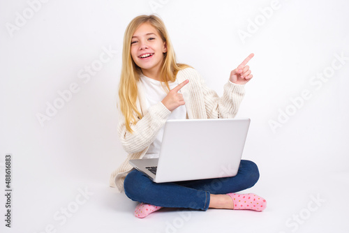 caucasian teen girl sitting with laptop in lotus position on white background with positive expression, indicates with fore finger at blank copy space for your promotional text or advertisement. © Jihan