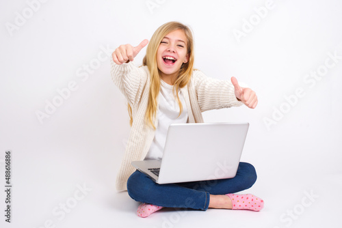 caucasian teen girl sitting with laptop in lotus position on white background making positive gesture with thumbs up smiling and happy for success. Looking at the camera, winner gesture. © Jihan