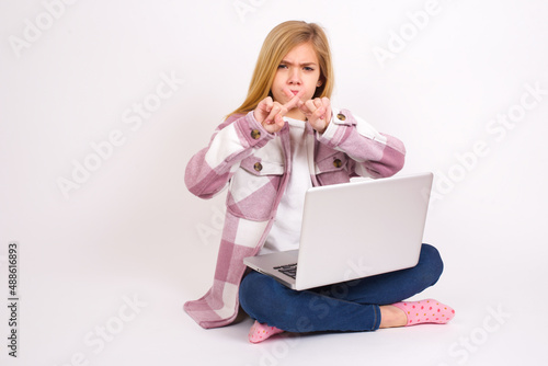 caucasian teen girl sitting with laptop in lotus position on white background has rejection angry expression crossing fingers doing negative sign. photo