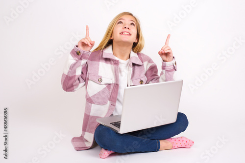 Successful friendly looking caucasian teen girl sitting with laptop in lotus position on white background exclaiming excitedly, pointing both index fingers up, indicating something. © Jihan
