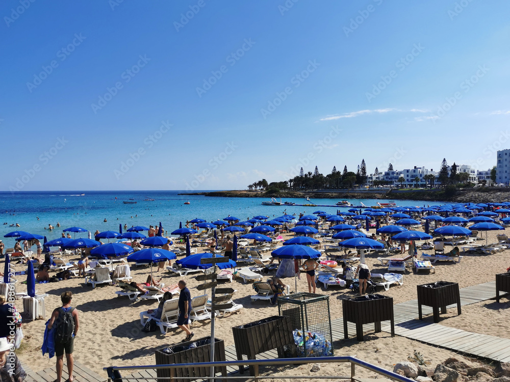 Protaras. Famagusta area. Cyprus. The sandy beach of Fig Tree Bay, people sunbathe and relax on sun loungers under umbrellas, swim in the Mediterranean Sea Protaras. Famagusta area. Cyprus. The sandy beach of Fig Tree Bay, people sunbathe and relax on sun loungers under umbrellas, swim in the Mediterranean Sea