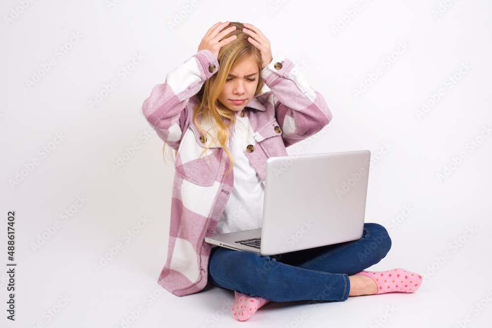 caucasian teen girl sitting with laptop in lotus position on white background holding head with hands, suffering from severe headache, pressing fingers to temples caucasian teen girl sitting with laptop in lotus position on white background holding head with hands, suffering from severe headache, pressing fingers to temples