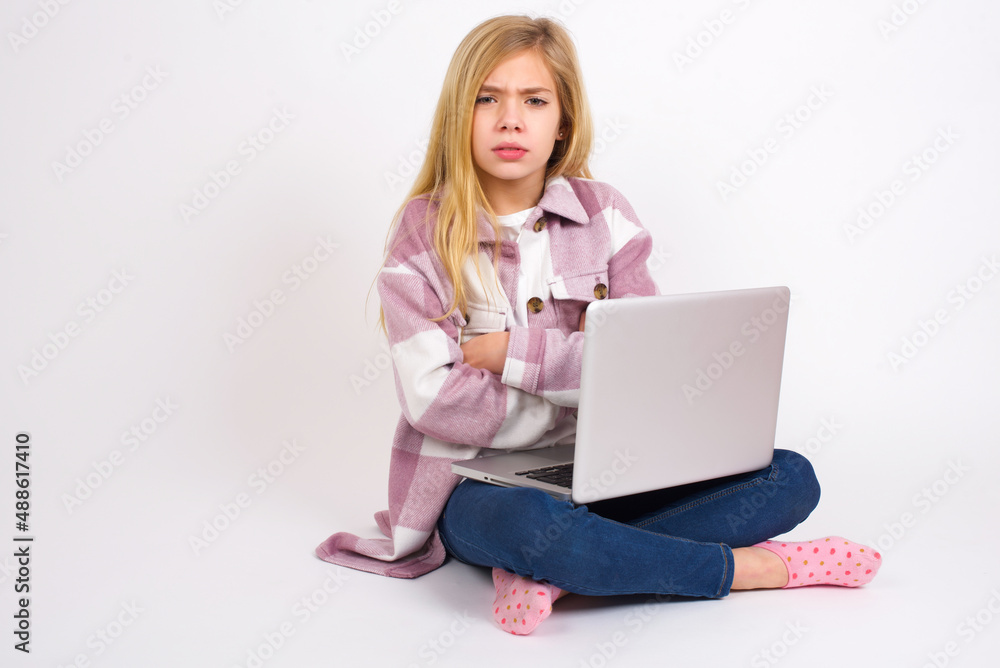 caucasian teen girl sitting with laptop in lotus position on white background frowning his face in displeasure, keeping arms folded, waiting for an explanation. caucasian teen girl sitting with laptop in lotus position on white background frowning his face in displeasure, keeping arms folded, waiting for an explanation.