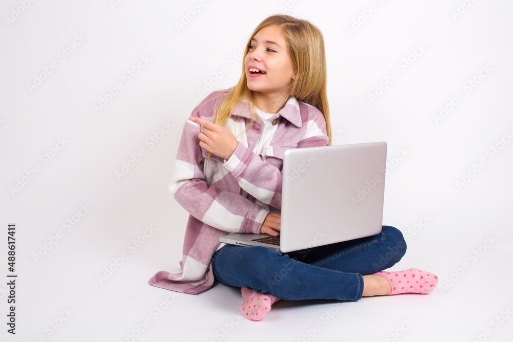 Happy cheerful smiling caucasian teen girl sitting with laptop in lotus position on white background looking and pointing aside with hand. Copy space and advertisement concept. Happy cheerful smiling caucasian teen girl sitting with laptop in lotus position on white background looking and pointing aside with hand. Copy space and advertisement concept.