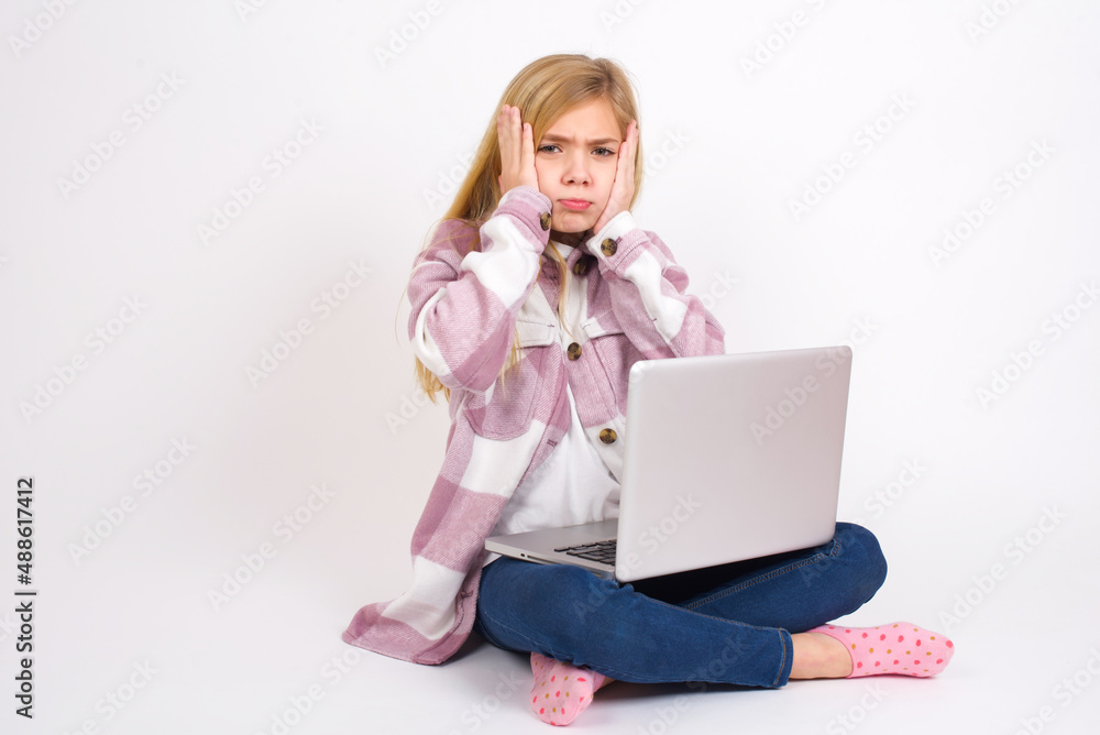 caucasian teen girl sitting with laptop in lotus position on white background holding head in hands with unhappy expression watching sad movie about animals and trying not to cry. caucasian teen girl sitting with laptop in lotus position on white background holding head in hands with unhappy expression watching sad movie about animals and trying not to cry.