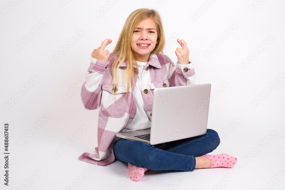 caucasian teen girl sitting with laptop in lotus position on white background holding fingers crossed with worried expression hoping boss didn't noticed mistakes at work. caucasian teen girl sitting with laptop in lotus position on white background holding fingers crossed with worried expression hoping boss didn't noticed mistakes at work.